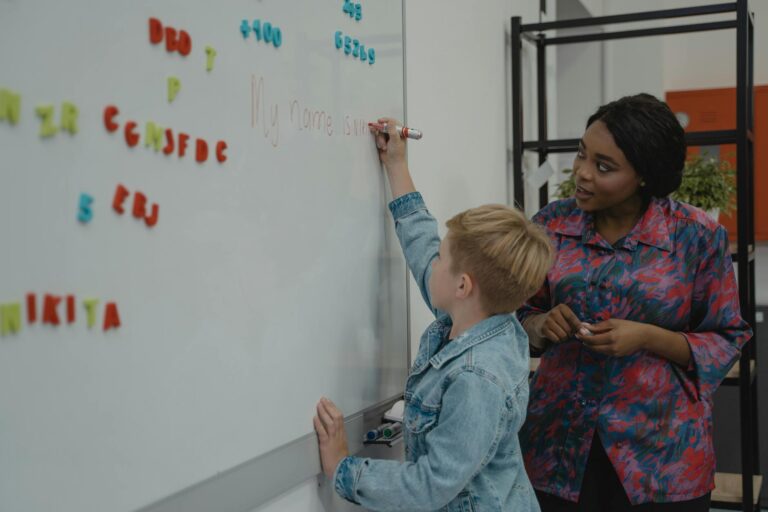 A teacher guides a student writing on a classroom whiteboard during a lesson, enhancing learning skills.